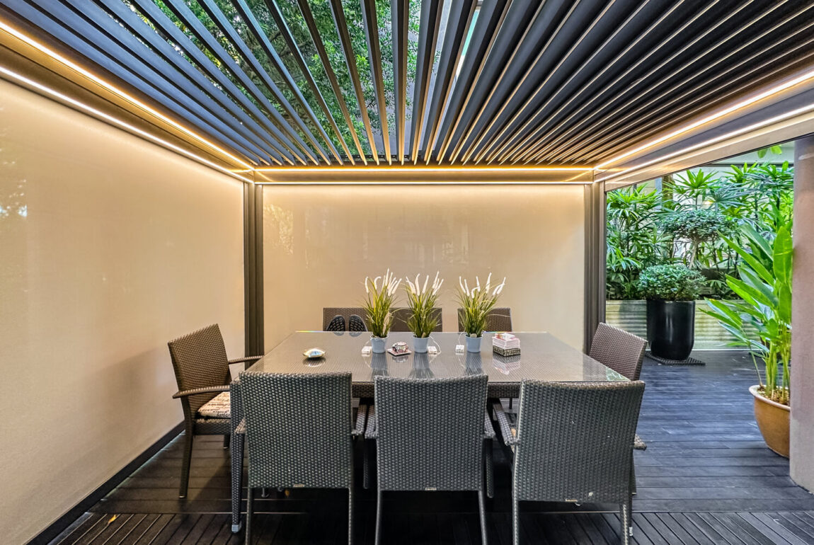 An outdoor structure made of sleek aluminium stands in a backyard. The structure features a flat roof with no canopy and is supported by sturdy pillars. The roof is comprised of horizontally-oriented louvers that can be controlled by an automated system to adjust the amount of sunlight and ventilation that enters the space. The louvers are angled to provide shade and privacy when closed, while also allowing for natural light and fresh air when opened. Zipblinds have been installed on the sides of the structure to provide additional shade and privacy, and can also be controlled by the automated system. The motorized system allows for easy operation of the louvers and zipblinds with the touch of a button. The pergola is surrounded by green plants and outdoor furniture, making it an inviting spot for outdoor relaxation and entertainment.