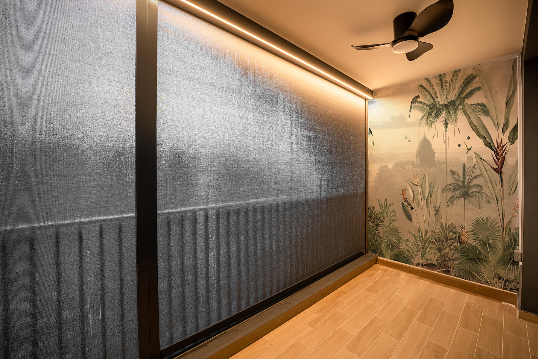 A balcony in Singapore featuring zip blinds, also known as zip track outdoor blinds, installed for renovation purposes. The blinds are partially closed, showing a tasteful tropical-themed mural on the adjacent wall, with palm trees, exotic birds, and a historical building in the background. Warm lighting from above casts a soft glow on the elegant wooden flooring and the sleek, modern ceiling fan adds a contemporary touch.