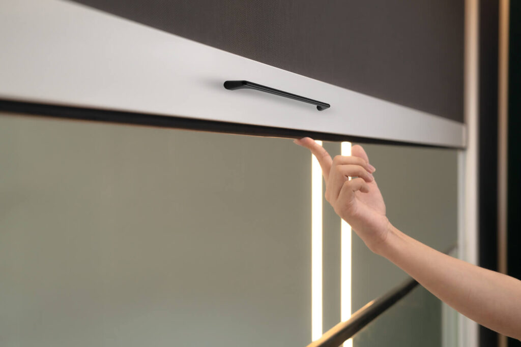 Close-up of a person’s hand gently pulling the black handle on the white bottom rail of a ZipBlind X track-guided outdoor blind, illustrating manual operation on a balcony